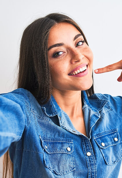 A young female wearing a denim button-down shirt and pointing to her smile after undergoing teeth whitening in Hoboken 