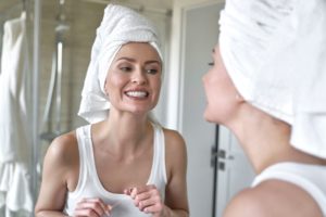 Woman in a white tank top in her bathroom with a white towel on her head looking at her teeth in the mirror holding floss